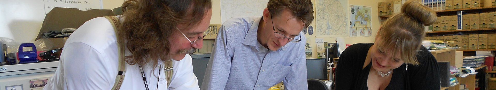 Archaeologists stand around a desk in an archive to look at maps.