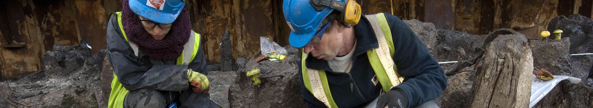 Two archaeologists in blue hard hats examine an archaeological feature.