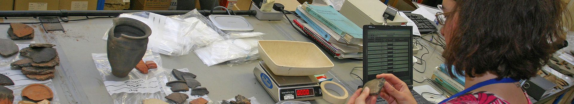 An archaeologist sits at a table with a computer, scales and pieces of ancient pottery.