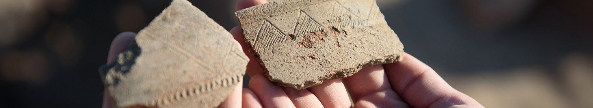 Hands hold two fragments of ancient pottery with incised decoration.