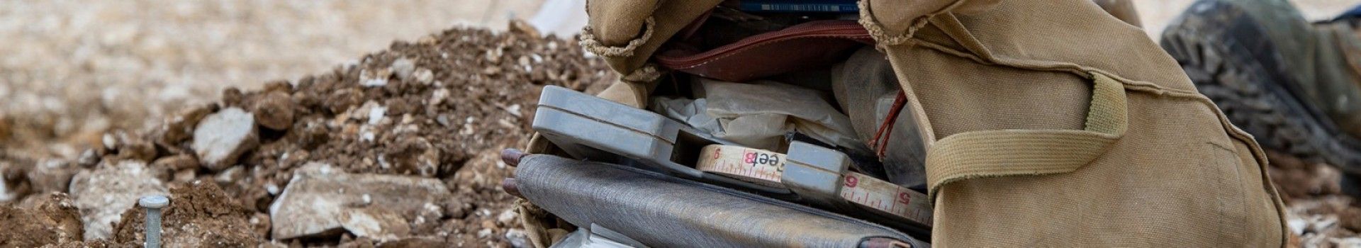 A satchel containing a measuring tape and notebook lies on a dusty ground.