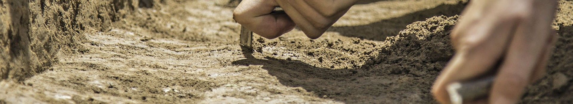 Hands holding trowels work in a trench on an archaeological excavation..