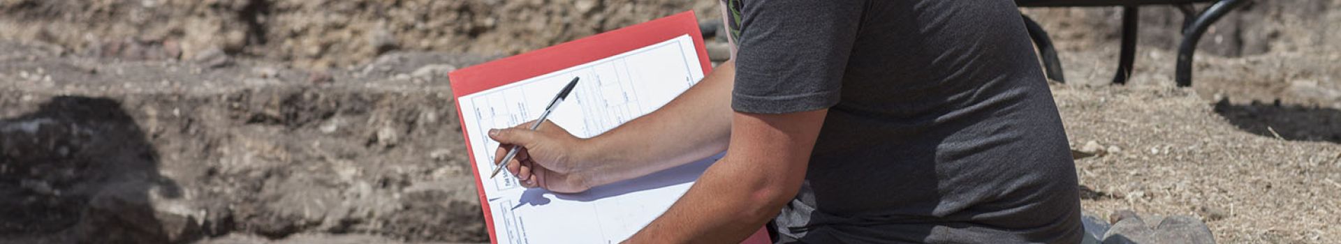 An archaeologist sits on the side of an excavation trench and completes context records.