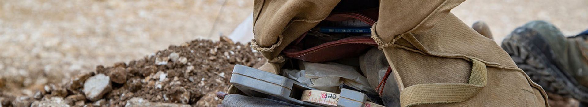 A satchel containing a measuring tape and notebook lies on a dusty ground.