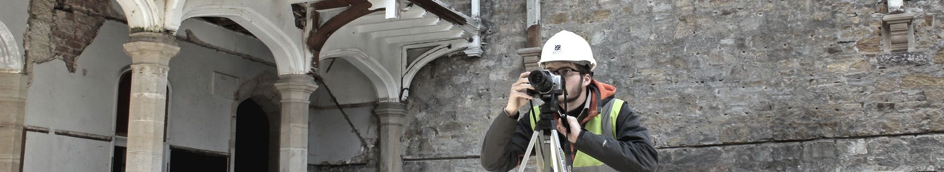 An archaeologist with a camera and tripod records an old, disused building.