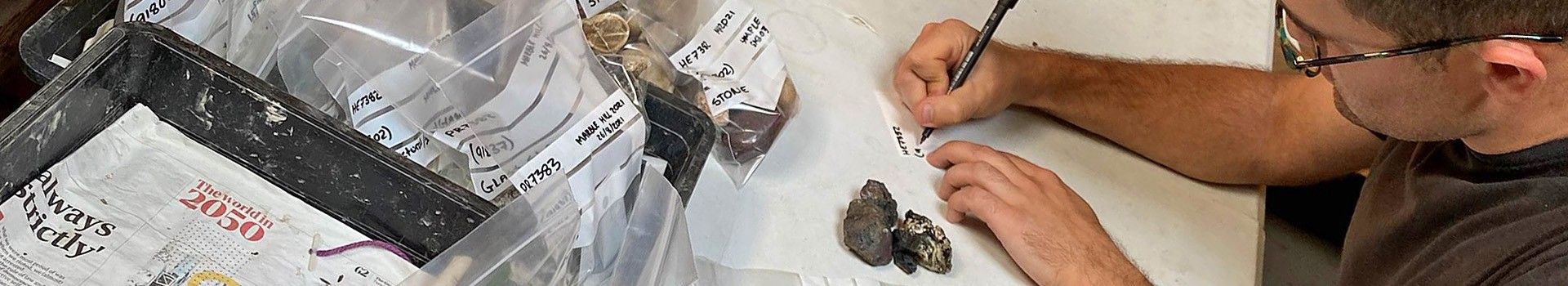 An archaeologist writes on clear plastic finds bags at a desk.