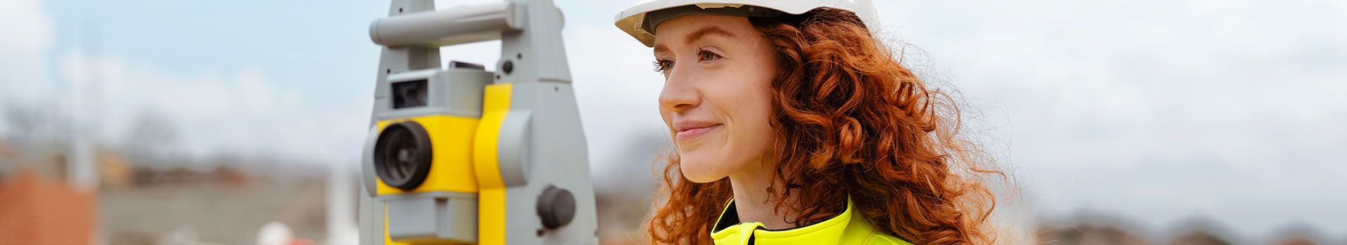 A female archaeologist in hi-vis clothing stands behind a surveying theodolite on a tripod.