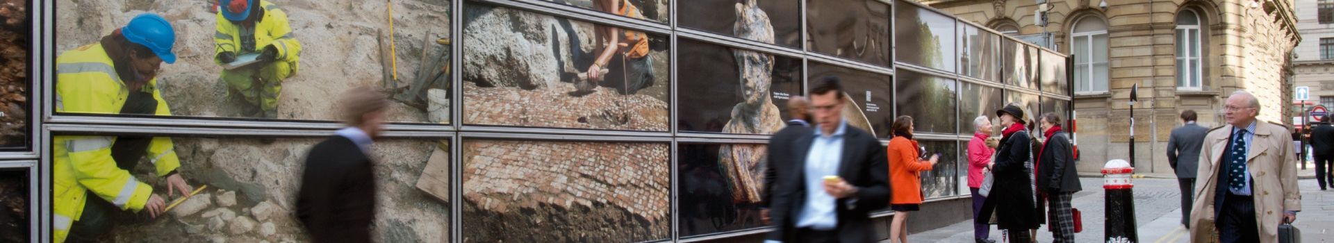 Pedestrians walk past street hoardings showing archaeological excavation photos.