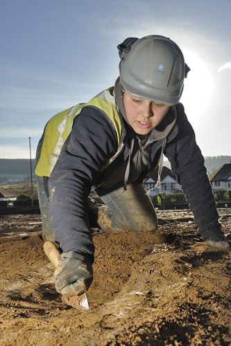 An archaeologist in a white hard hat holds a trowel and excavates.