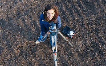 An archaeologist stands next to a surveying tripod and looks up at the camera.