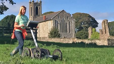 An archaeologist undertakes geophysical survey in a field with a church in the background.