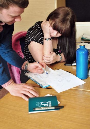 A woman sits at a table to listen to a tutor, who points at pieces of paper.