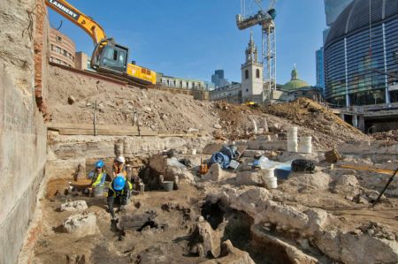 Archaeologists excavate in the middle of an urban area surrounded by construction equipment.