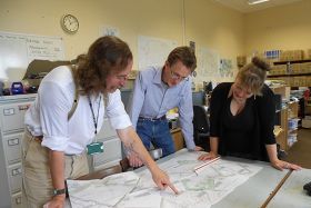 Archaeologists stand around a large desk and examine maps.