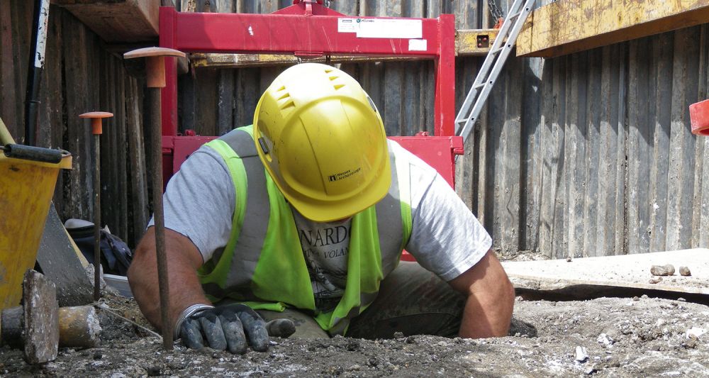 An archaeologist in a hard hat kneels to excavate a feature in a deep hole.