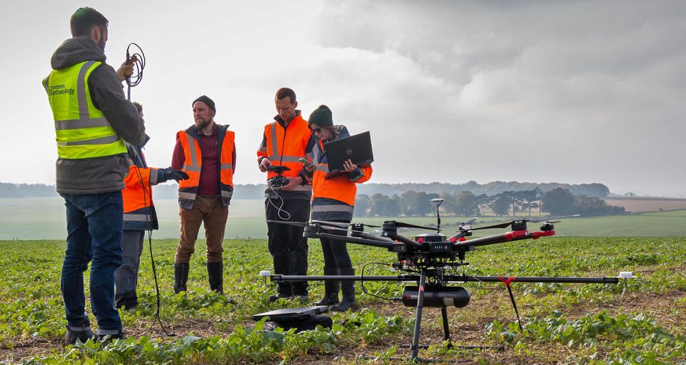 Archaeologists stand next to a drone and plan a flight in a green landscape.