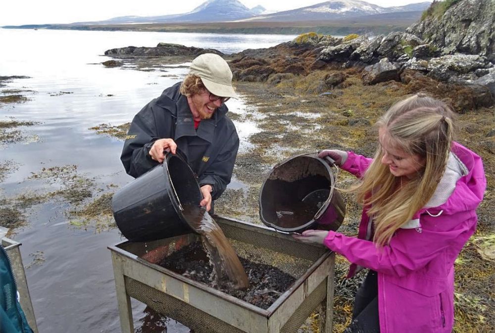 Two young archaeologists pour buckets into a sieve in a rocky coastal landscape.