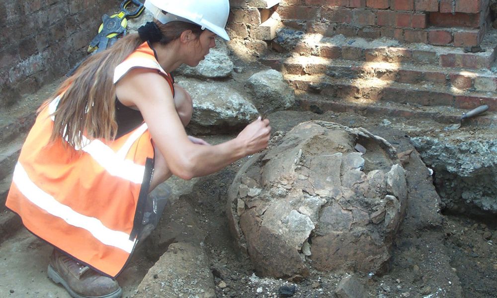 A young archaeologist in orange hi-vis clothing excavates a large pottery vessel.