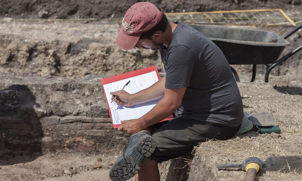 An archaeologist sits on the side of an excavation trench and completes context records.