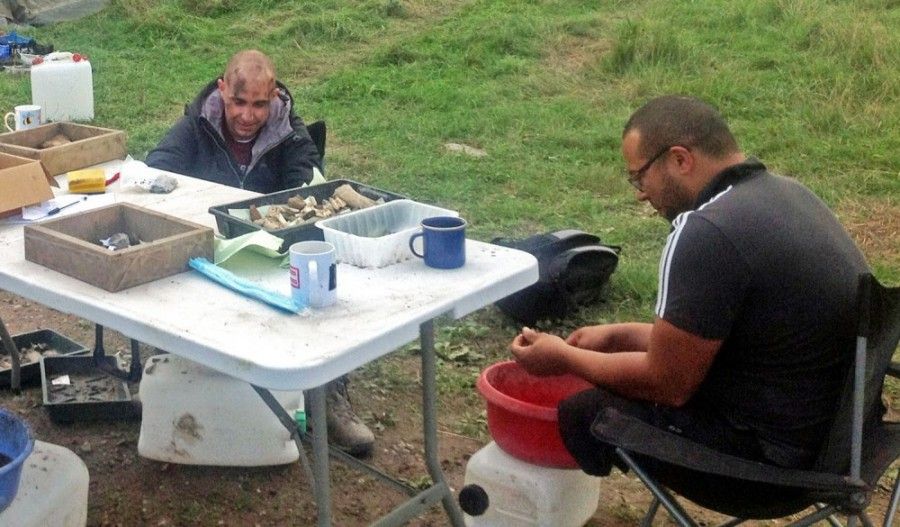 Two partipants in Operation Nightingale sit at a table outside and wash pottery finds.