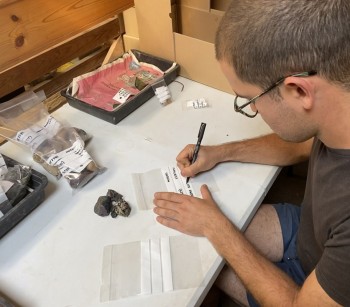 An archaeologist writes on clear plastic finds bags at a desk.