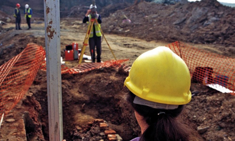 An archaeologist holds a levelling staff whilst a colleague surveys an excavation site.