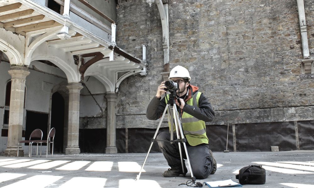 An archaeologist with a camera and tripod records an old, disused building.