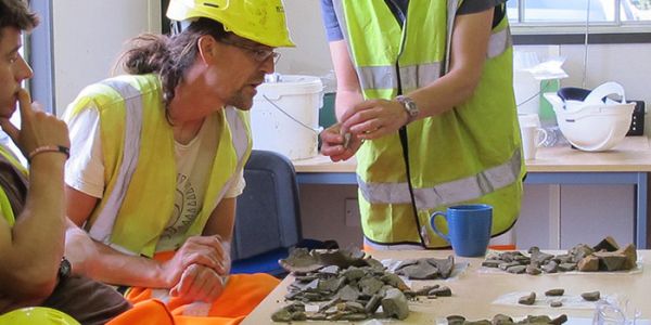 Archaeologists listen to a finds expert who displays recovered finds.