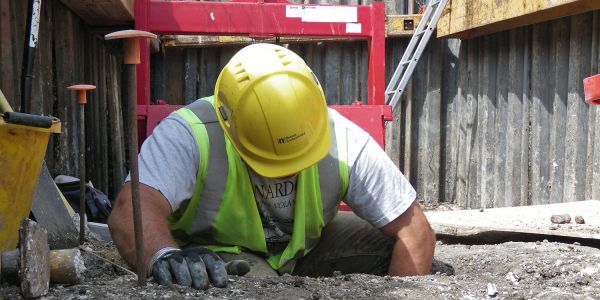 An archaeologist in a hard hat kneels to excavate a feature in a deep hole.