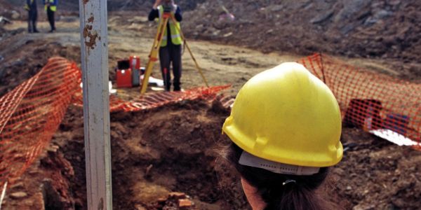 An archaeologist holds a levelling staff whilst a colleague surveys an excavation site.