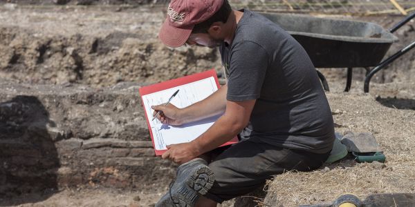 An archaeologist sits on the side of an excavation trench and completes context records.