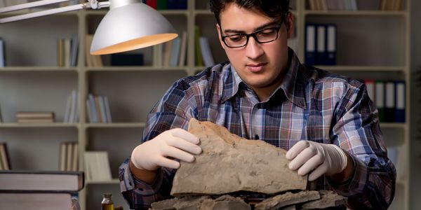 An archaeologist wears latex gloves and examines rock samples under a desk-lamp.