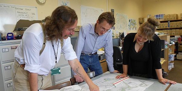 Archaeologists stand around a large desk and examine maps.