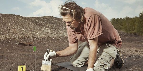 A young archaeologist with a brush kneels on an archaeological site.