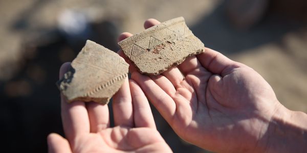 Hands hold two fragments of ancient pottery with incised decoration.