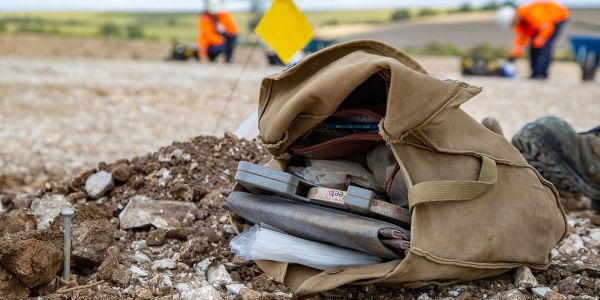 A satchel containing a measuring tape and notebook lies on a dusty ground.