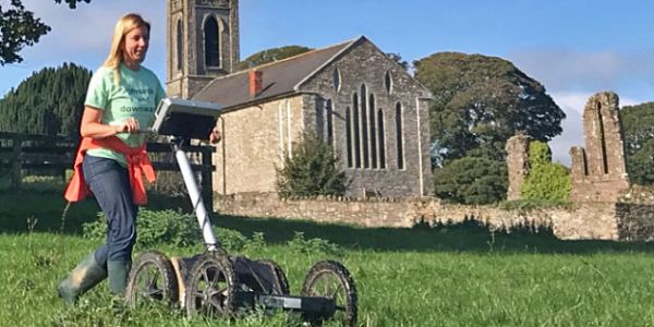 An archaeologist undertakes geophysical survey in a field with a church in the background.