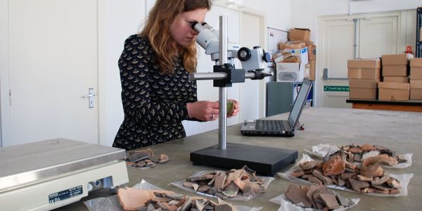 An archaeologist surrounded by piles of pottery examines an ancient sherd through a microscope.