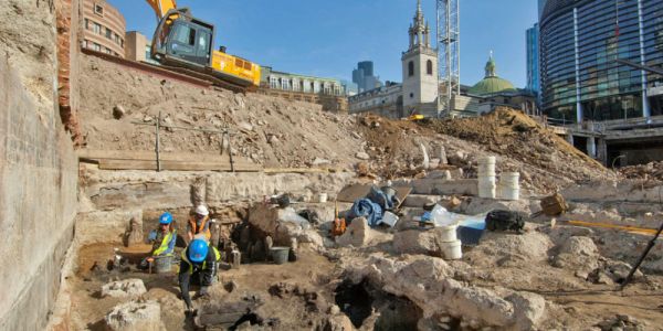 Archaeologists excavate in the middle of an urban area surrounded by construction equipment.